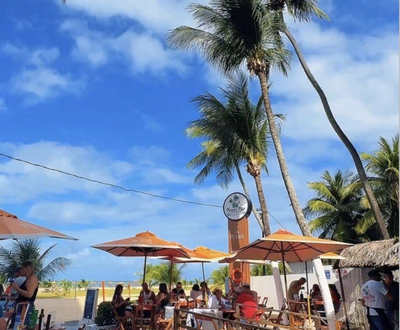 Área externa do Fruta Pão com guarda-sóis laranja, coqueiros altos e a faixa de areia ao fundo