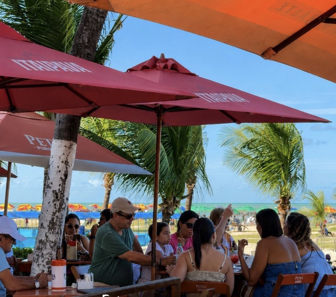 Mesas ocupadas sob guarda-sóis coloridos, com coqueiros e a praia de Candeias ao fundo