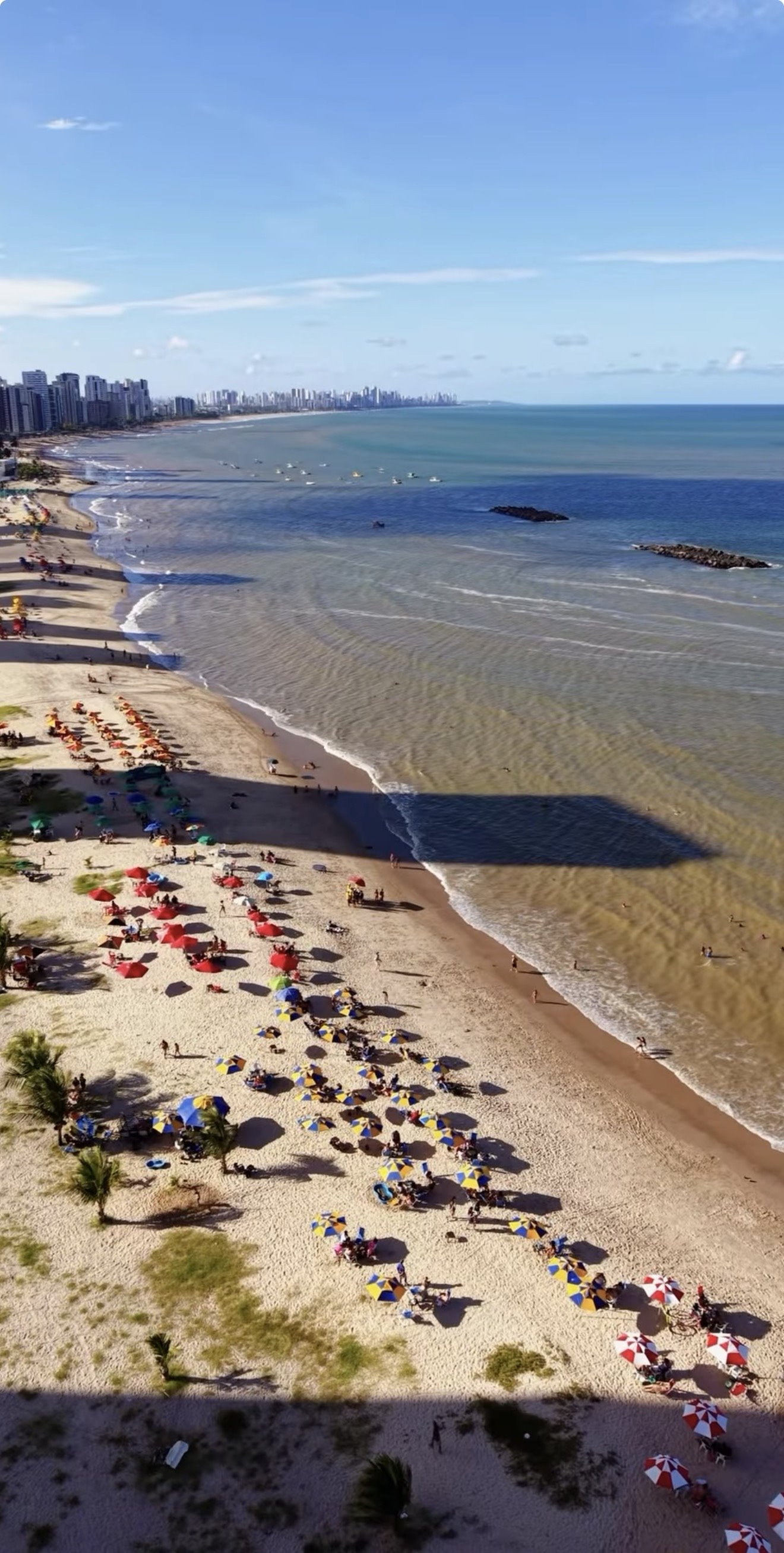 Vista aérea da praia de Candeias: mar azul, faixa de areia com guarda-sóis coloridos e a orla de Jaboatão ao fundo