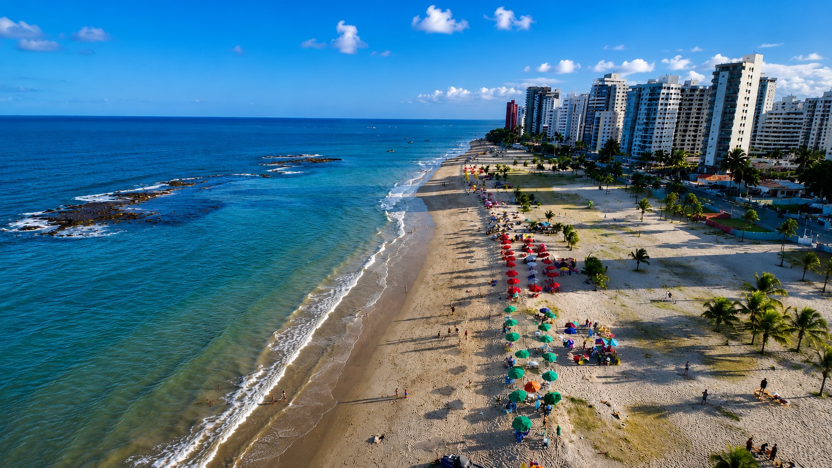 Vista aérea da praia de Candeias: mar azul com recife, ondas, faixa de areia e guarda-sóis coloridos